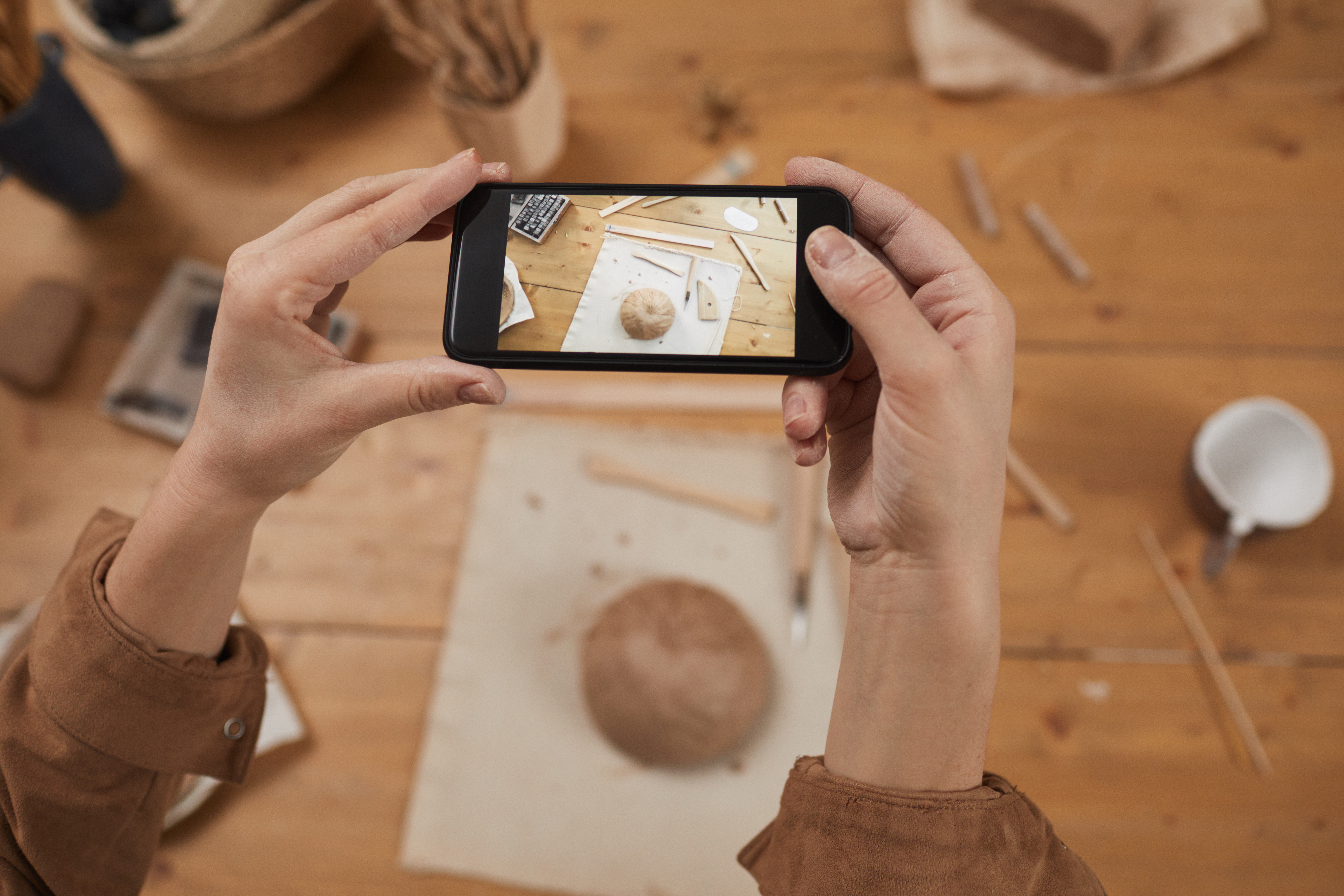 High angle close up of young woman taking smartphone photo of raw ceramic bowl in pottery workshop, small business concept, copy space
