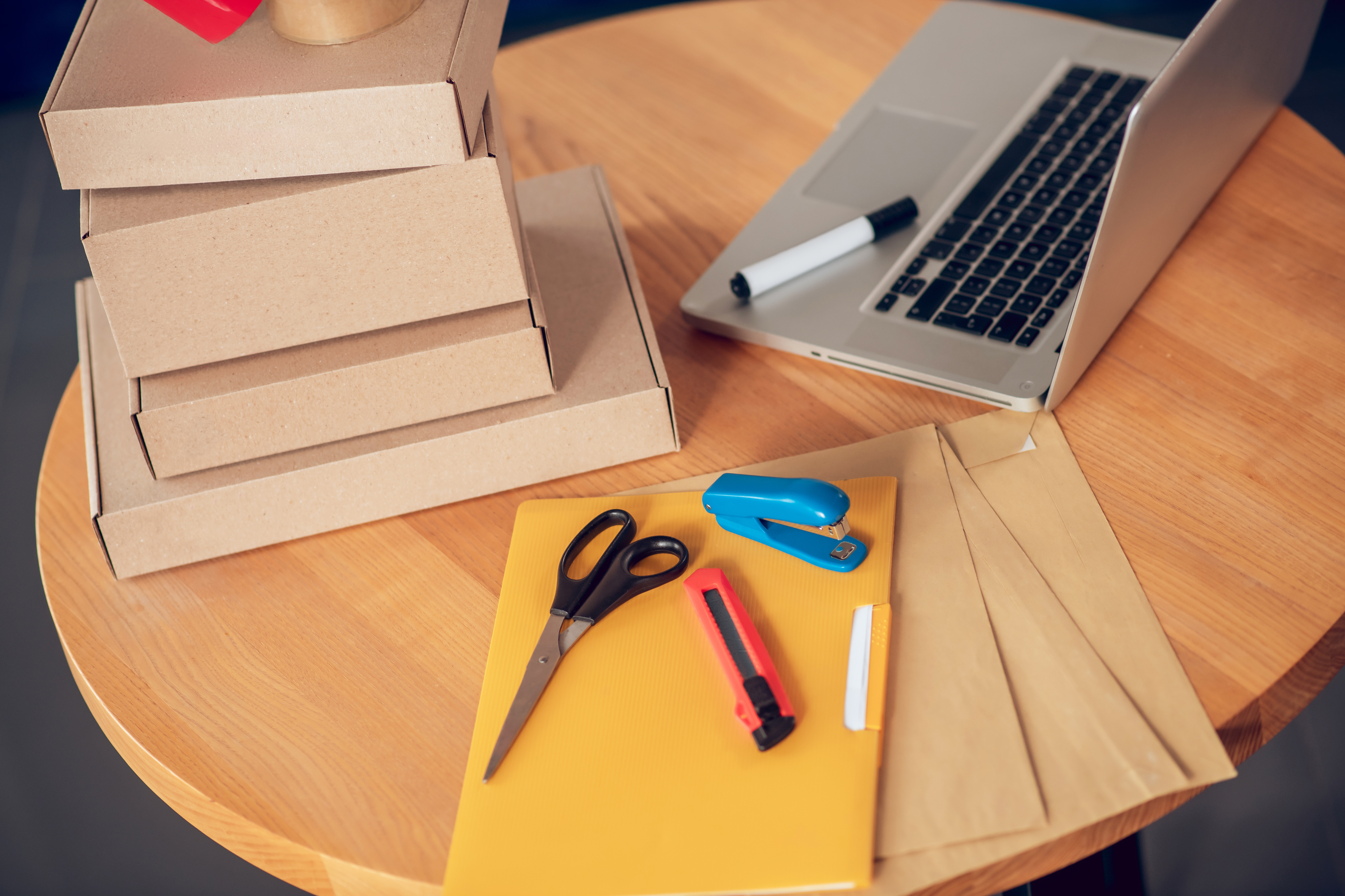 Laptop placed on a round wooden desk among stacked cardboard boxes and other office supplies